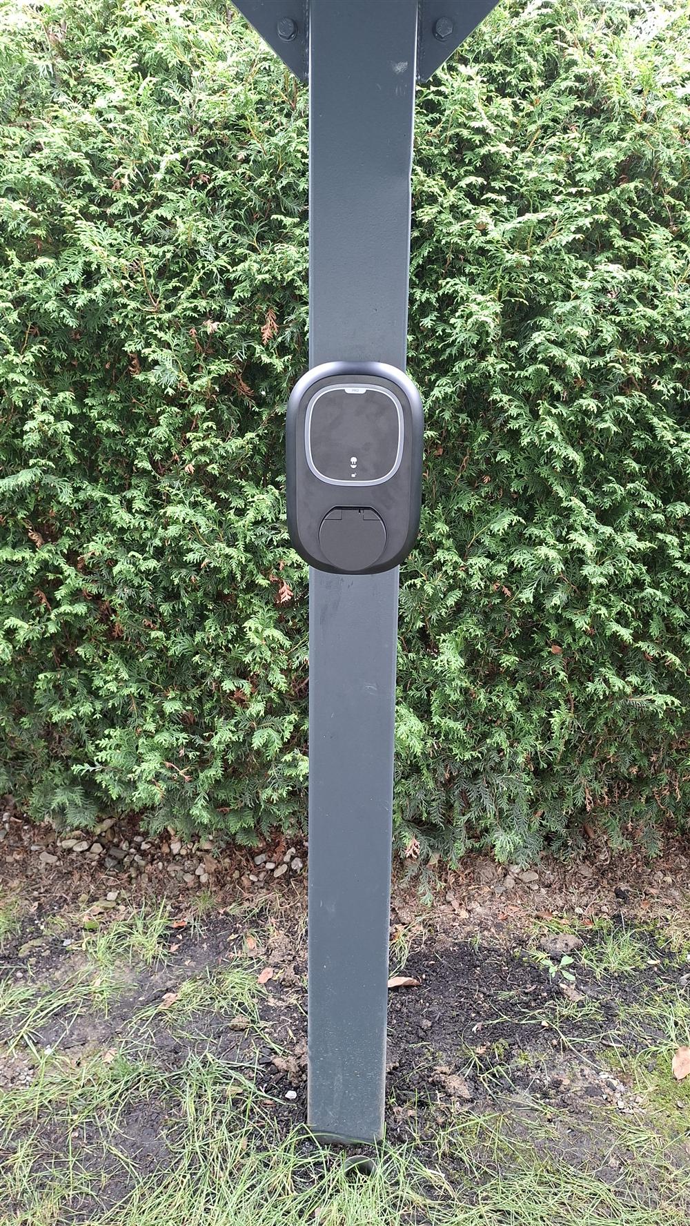 Tmavý wallbox namontovaný na samostatném sloupku u živého plotu